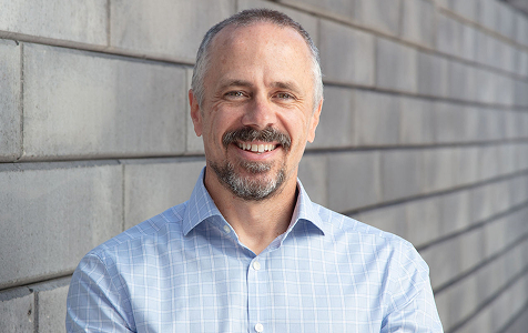 Smiling middle-aged man with short gray hair and a mustache and beard, wearing a light blue checkered shirt, standing in front of a gray brick wall.