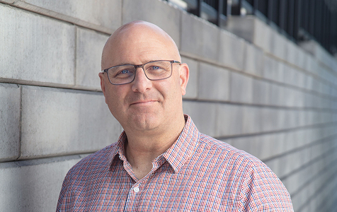 Middle-aged bald man wearing glasses and a checkered shirt standing in front of a concrete block wall.