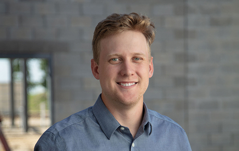 Smiling young man with short light brown hair wearing a blue button-up shirt standing indoors in front of a blurred gray brick wall and window.