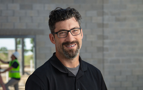 Smiling man with short curly dark hair, glasses, and a beard, wearing a black collared shirt, standing indoors with unfinished stone walls and a blurred person working in the background.