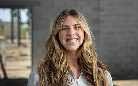 Smiling young woman with long wavy blonde hair wearing a light gray zip-up top against a blurred background of a concrete wall and outdoor area.