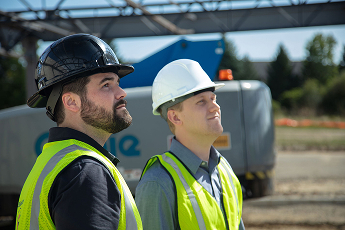 Two construction workers wearing hard hats and yellow safety vests looking up at a construction site with machinery in the background.