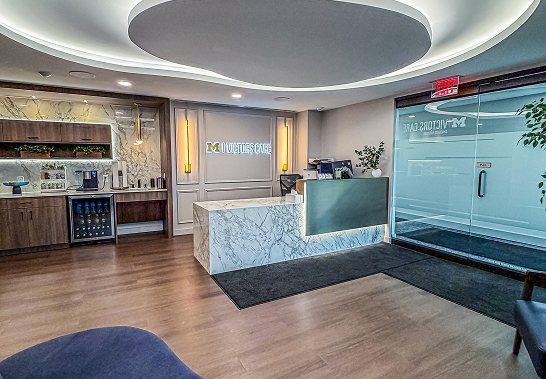 Modern reception area with a marble front desk and illuminated sign reading 'M VICTORS CARE' on the back wall, wooden flooring, glass door entrance, and built-in shelving with a small refrigerator and coffee station.