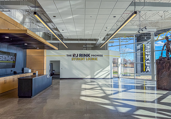 Bright modern student lounge with polished concrete floor, reception desk on the left with SEMCA logo, large windows on the right letting in natural light and casting shadows, and a wall with the text 'THE RJ RINK PROMISE STUDENT LOUNGE'.
