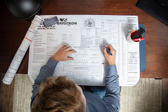 Person sitting at desk reviewing and marking a detailed engineering blueprint, with rolled-up plans and a red cup holding pens nearby.