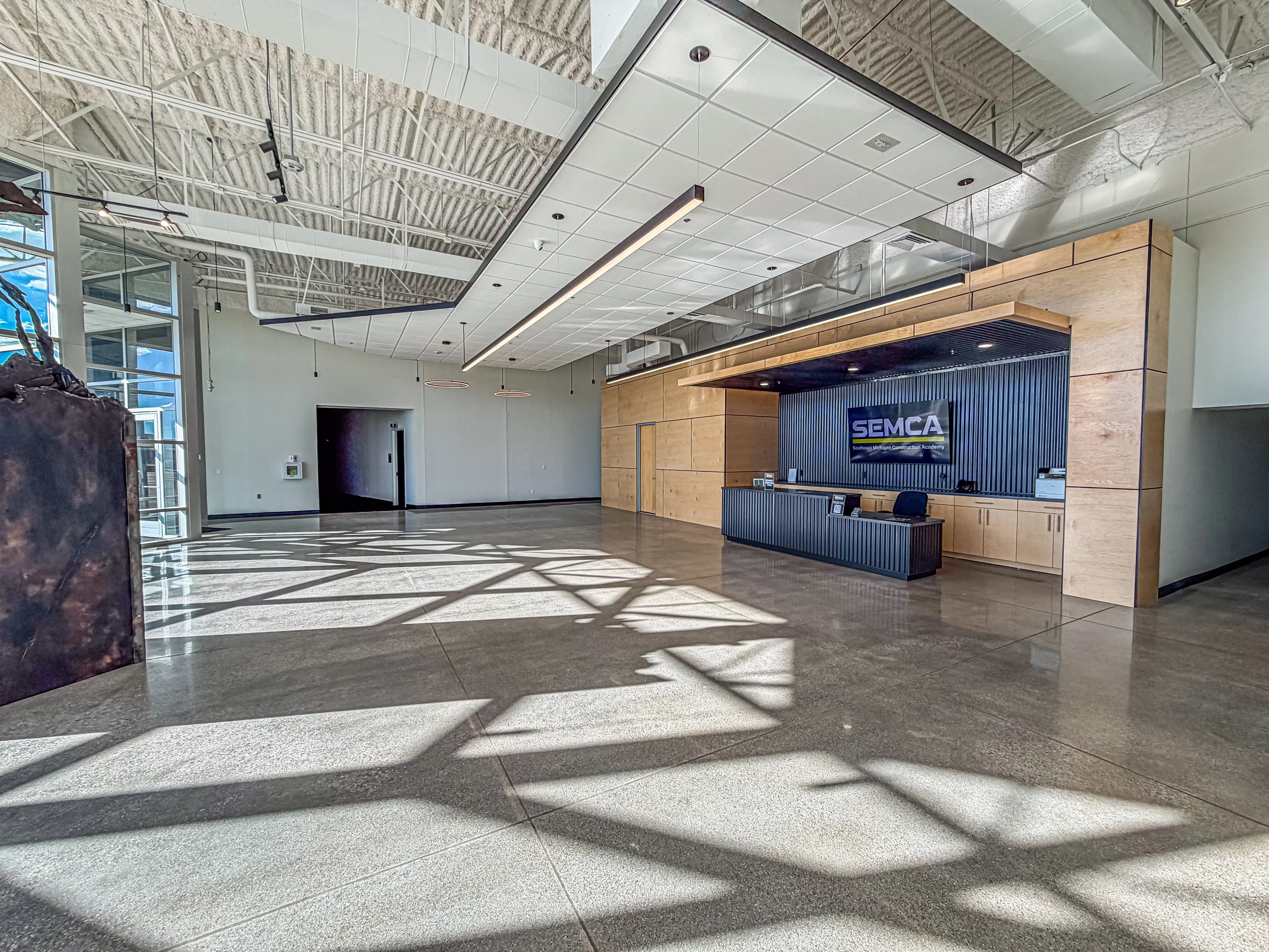 Bright, spacious modern lobby with polished concrete floors and sunlight casting geometric shadows through large left-side windows, featuring a wood-paneled reception area with a striped black counter and a sign reading 'SEMCA' on the back wall.