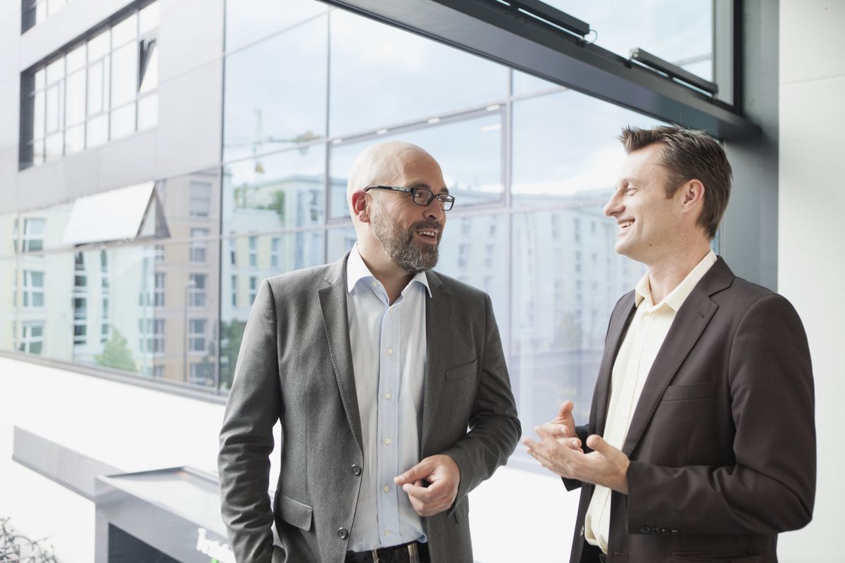 Two men in jackets talking and smiling in front of a glass-fronted office building