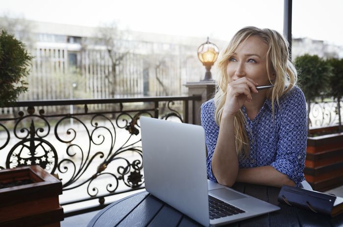 Woman sitting at an outdoor table with a laptop, pen at her lips, looking away in thought