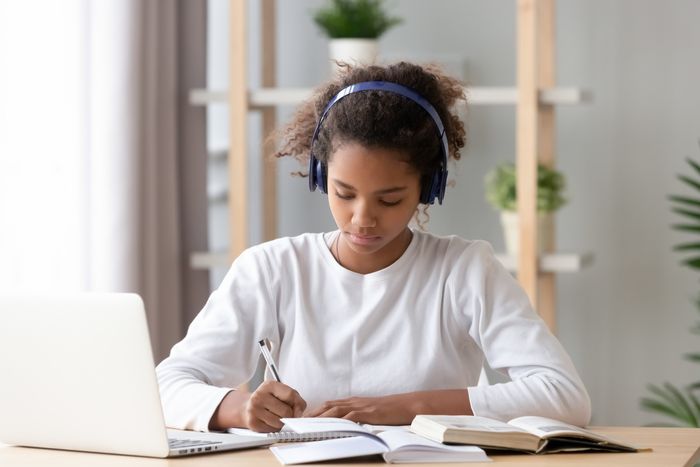 A young woman wearing headphones writes in a notebook at a desk beside a laptop