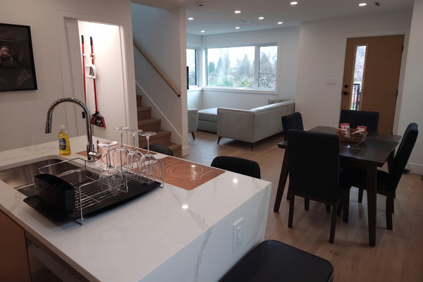 Modern open-concept living space with white marble kitchen island featuring a drying rack with wine glasses, a stainless steel sink, and dish soap, adjacent to a dining table with four black chairs and a bowl of snacks, with a white sectional sofa near large windows and a wooden staircase in the background.