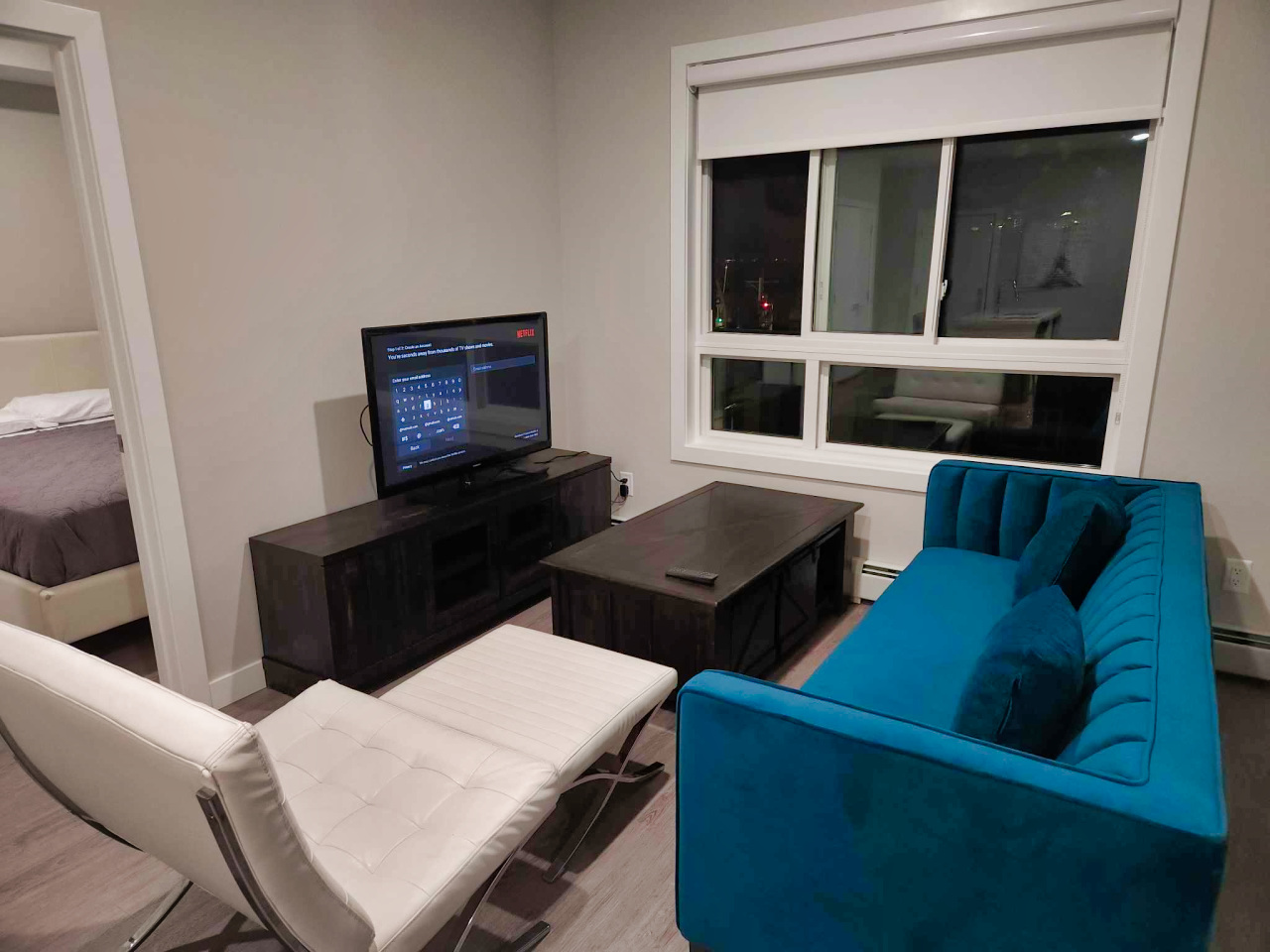 Living room with a blue velvet sofa with matching pillows, a white cushioned lounge chair, a wooden coffee table with a remote on it, and a dark wooden TV stand with a flat-screen TV displaying a Netflix screen for account login, next to a large window with a white roller shade. A bedroom with a bed is partially visible through an open door.