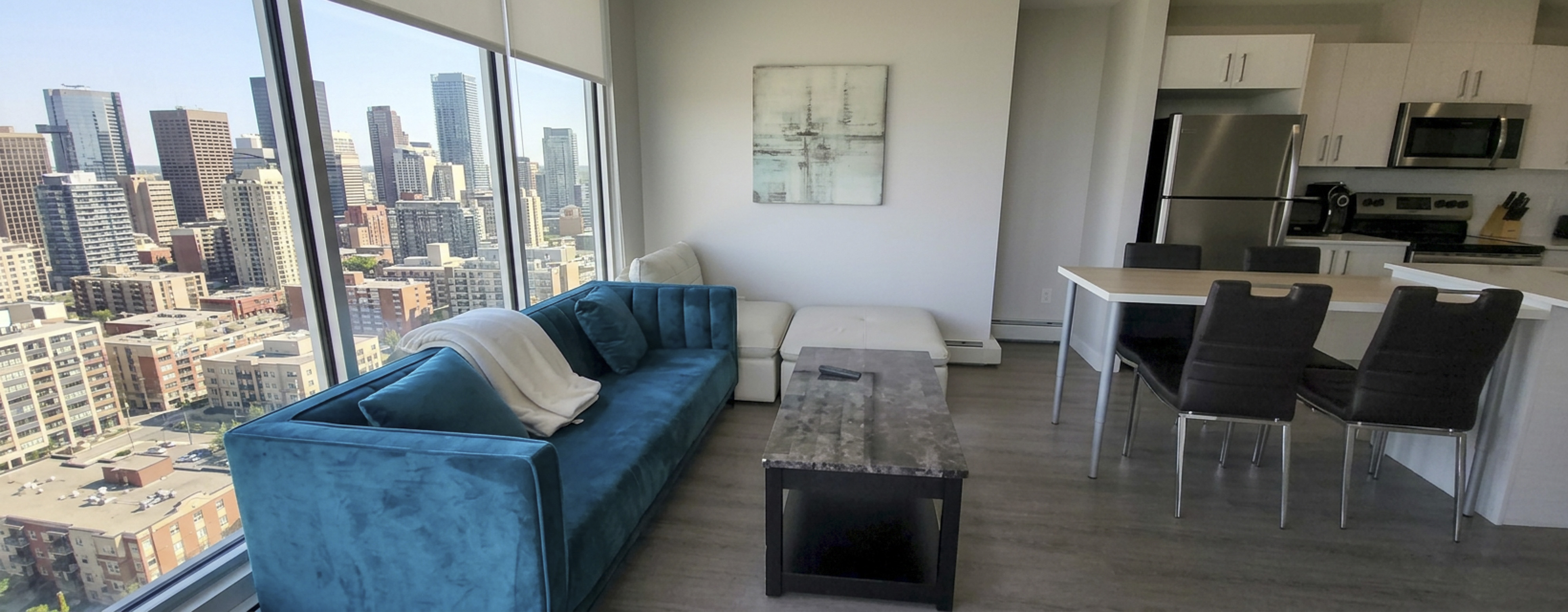 Modern apartment living room with a large turquoise sofa, white ottoman, dark coffee table, dining table with four black chairs, and a kitchen area with stainless steel appliances, set against floor-to-ceiling windows showing a city skyline.