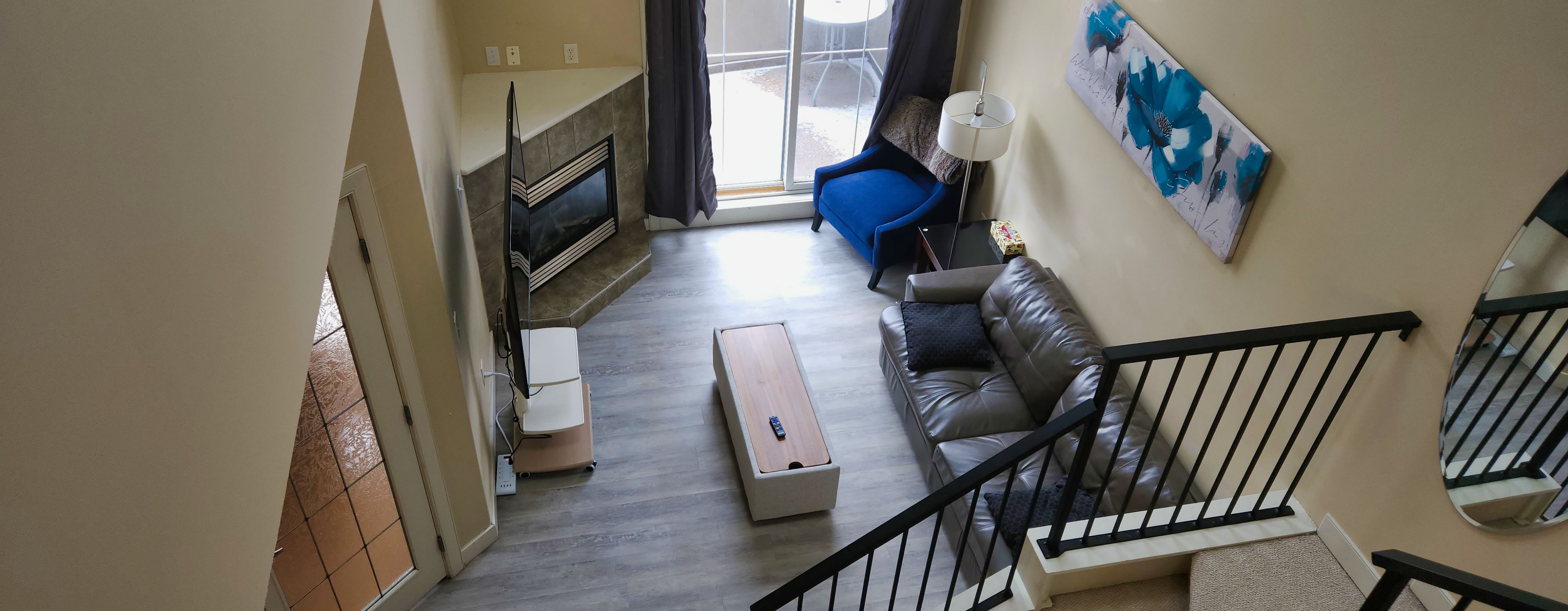 Modern living room viewed from upstairs, featuring a gray leather sofa with two black pillows, a blue armchair, a wooden coffee table with a remote, a side table with a lamp and tissue box, a TV on a stand, a corner fireplace with tile surround, and sliding glass doors with dark curtains leading outside. Beige walls, light wood flooring, and a staircase with black metal railing.