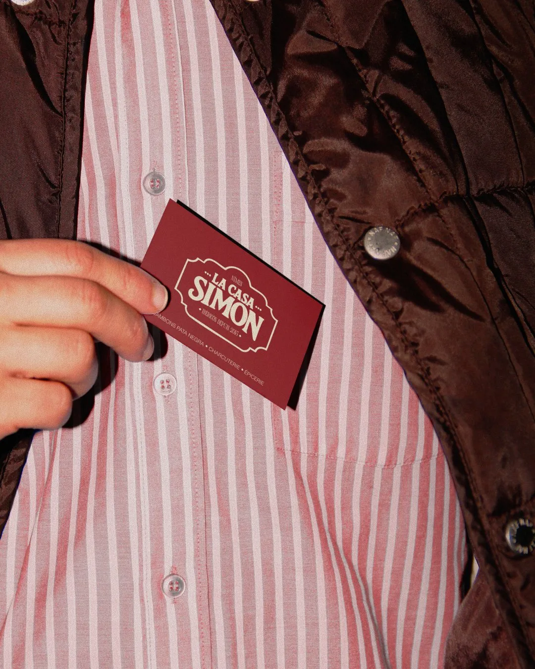 Main tenant une carte de visite bordeaux de La Casa Simon contre une chemise rayée rose, sous une veste brune matelassée.