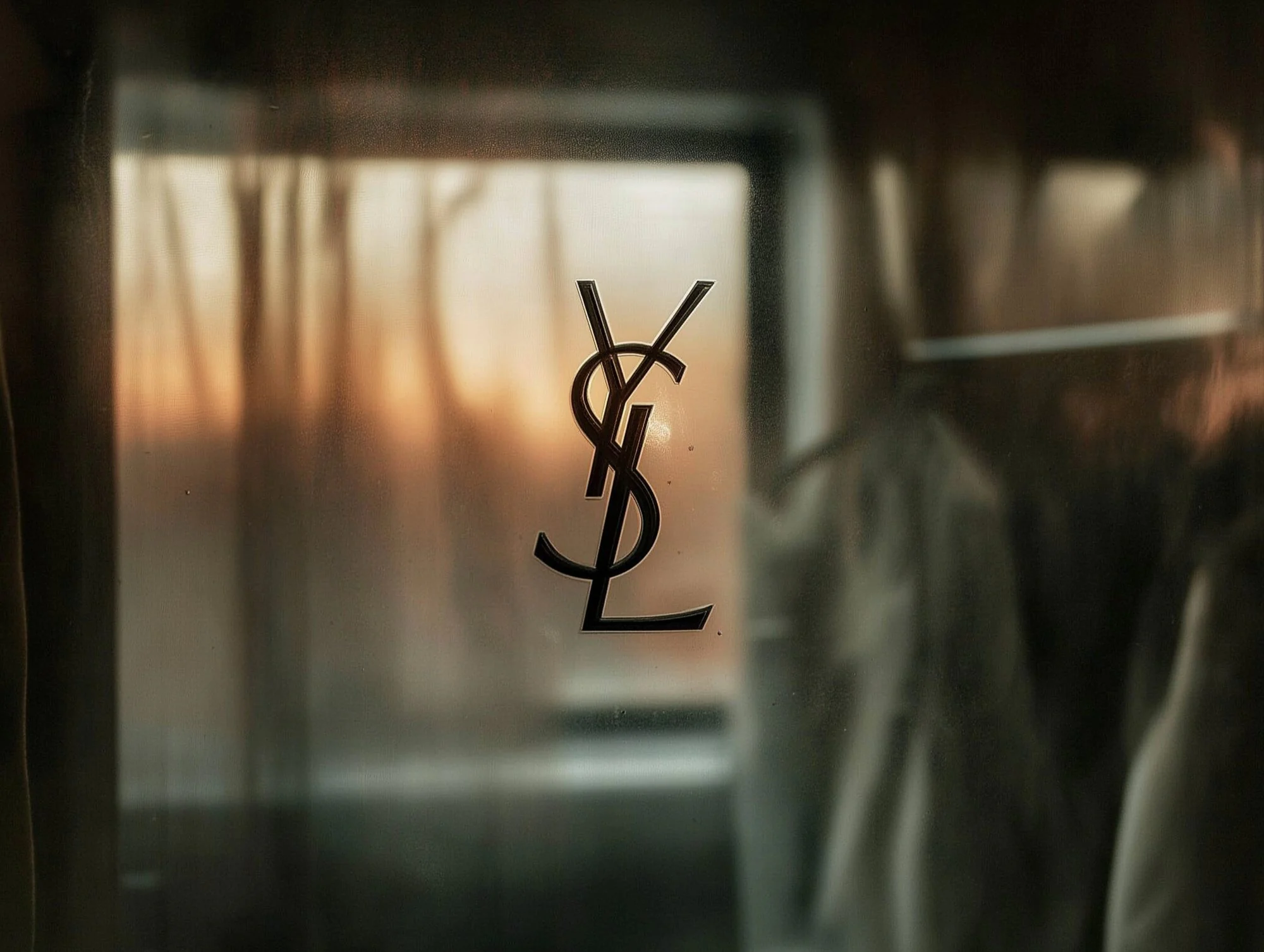 Monogramme YSL noir sur une vitre embuée, devant une fenêtre au couchant et des vêtements suspendus, flous.
