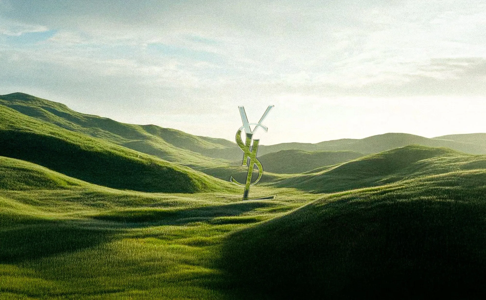 Monogramme YSL argenté planté au milieu de collines couvertes d’herbe verte, sous un ciel clair.