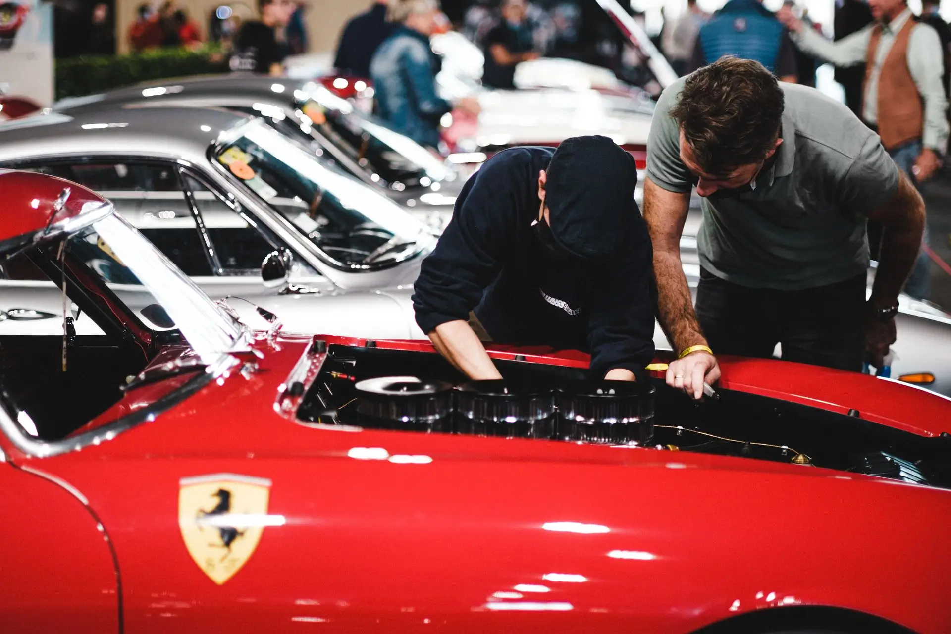 Two men closely inspecting or repairing the engine of a red Ferrari sports car with its hood open at a car show, with several vintage cars and other people visible in the background.