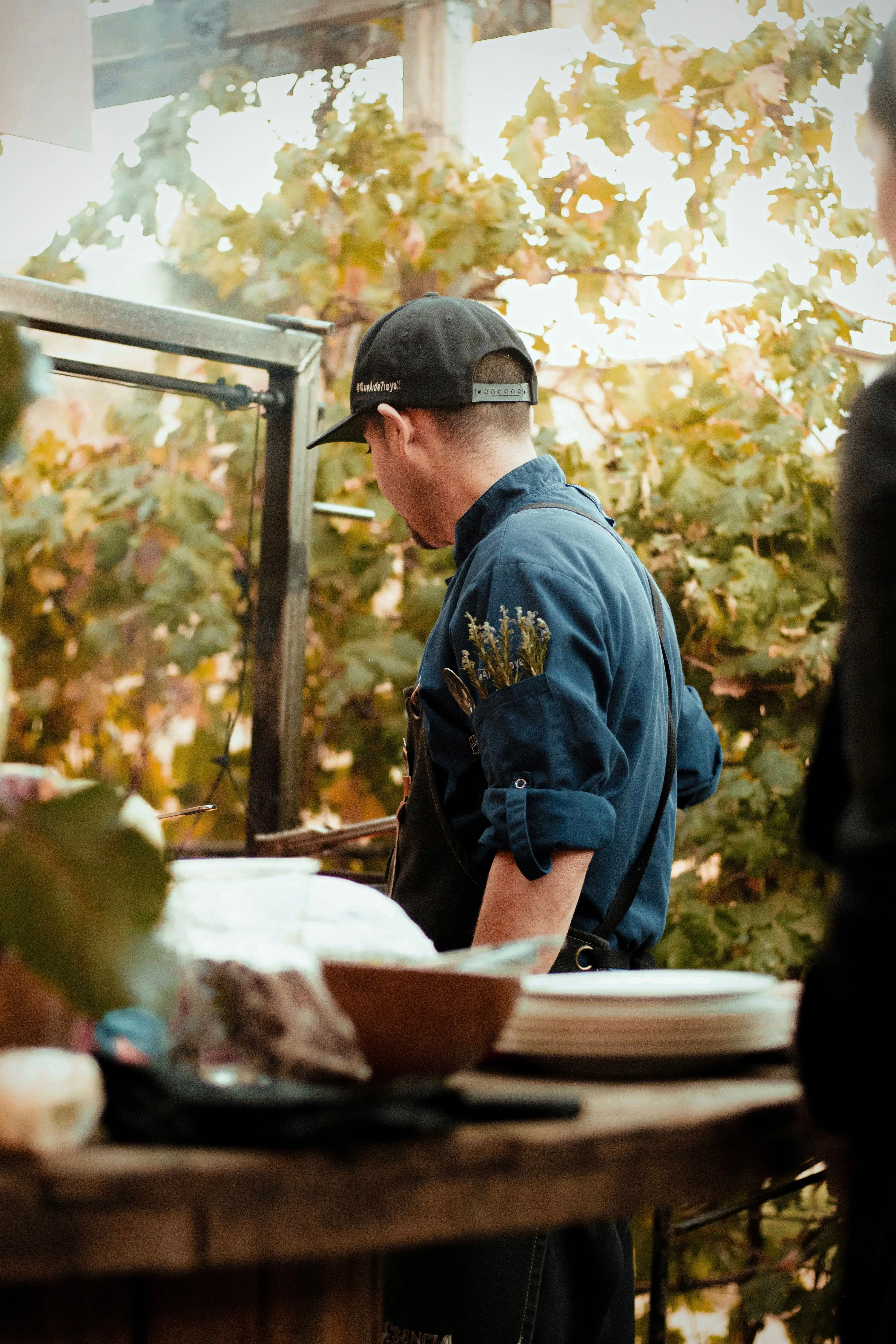 Man wearing a black cap and dark apron with herbs in the sleeve pocket, standing outdoors near a wooden table with plates and bowls, surrounded by green foliage.