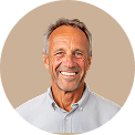 Smiling middle-aged man with short gray hair wearing a light blue collared shirt against a beige background.