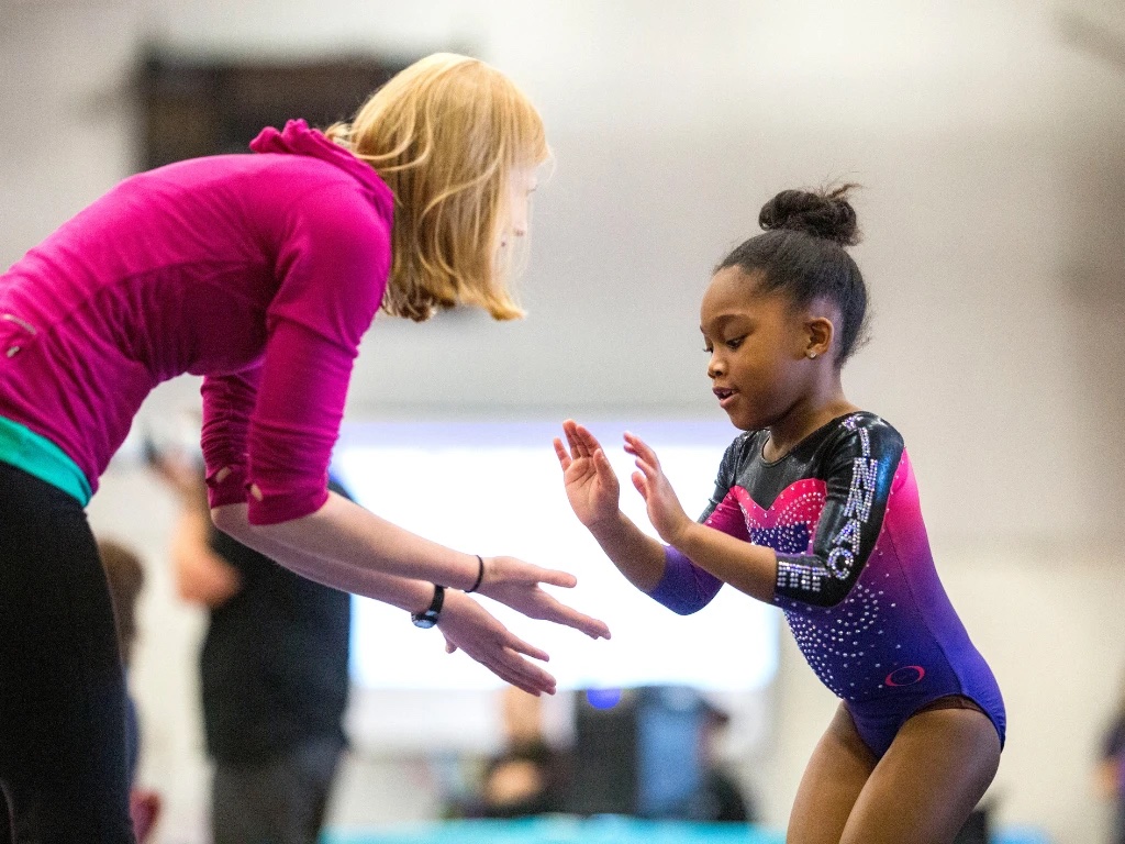A coach guiding a young gymnast through a skill in a club gym