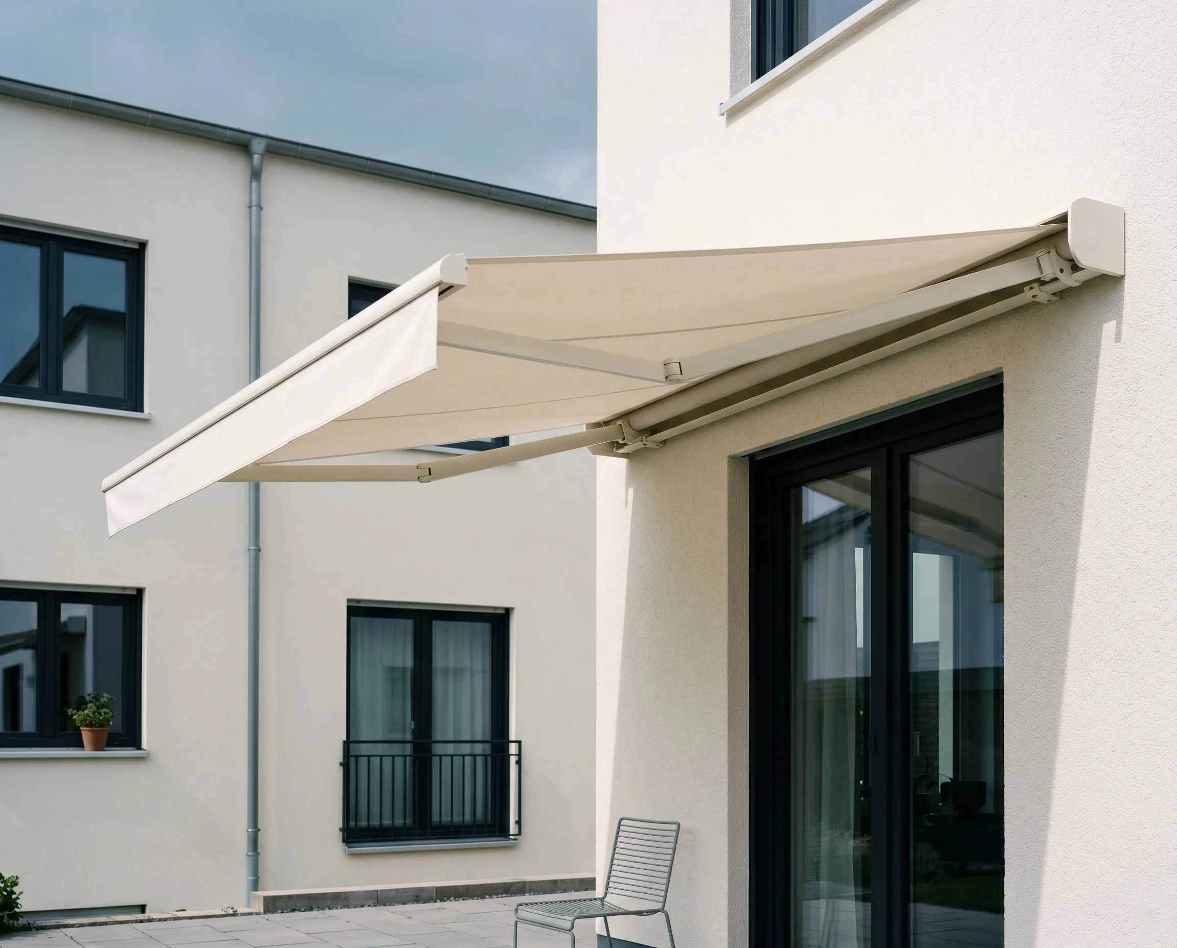 Hellbeige Markise ausgefahren am weißen Haus über einer Terrasse mit einem Metallstuhl.