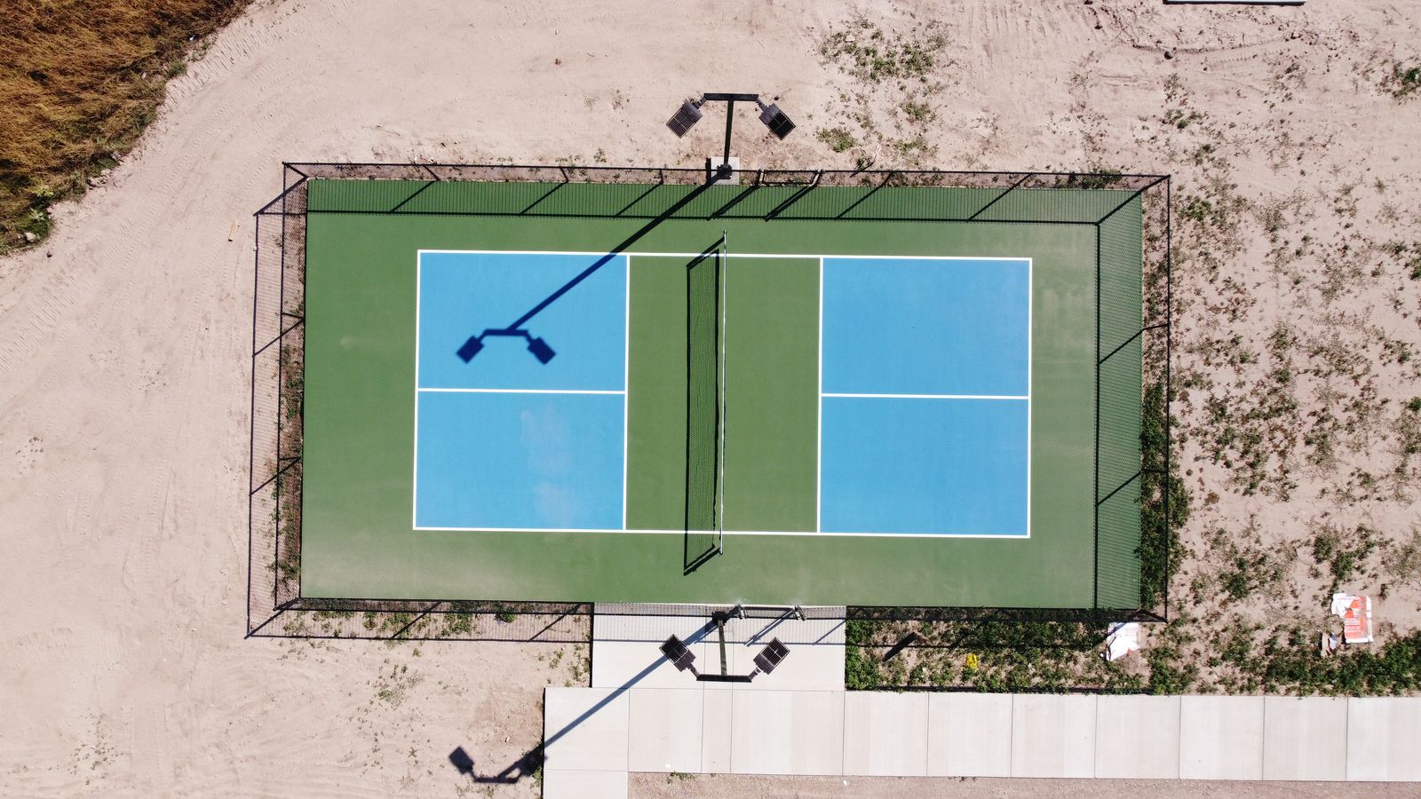 Aerial view of a fenced blue and green pickleball court with lighting poles on opposite sides.