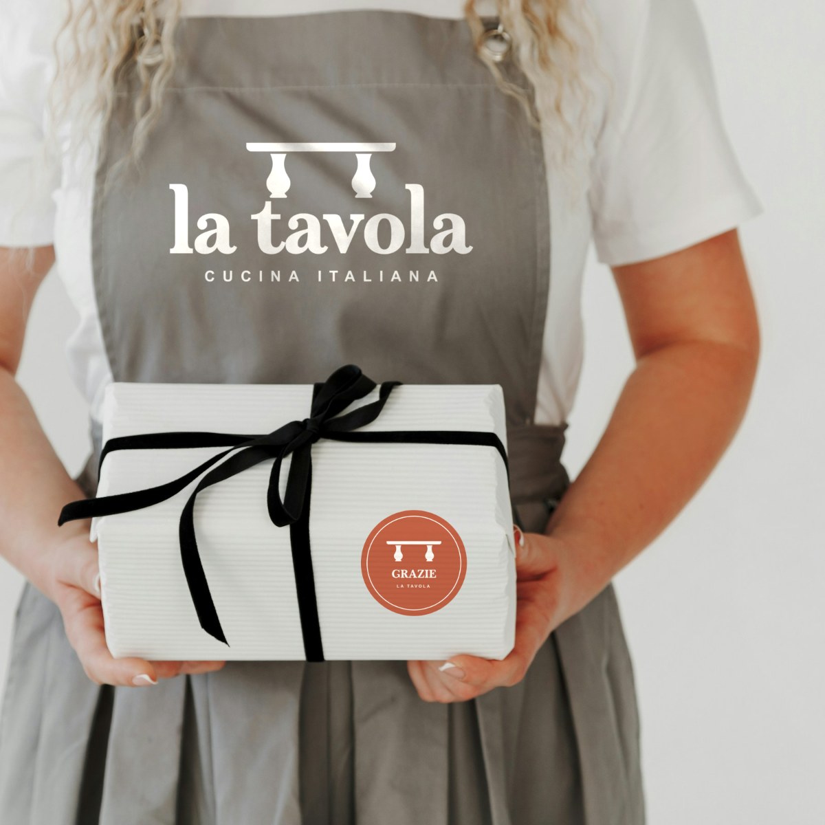 Staff apron printed with the La Tavola logo, holding a gift box with a Grazie seal