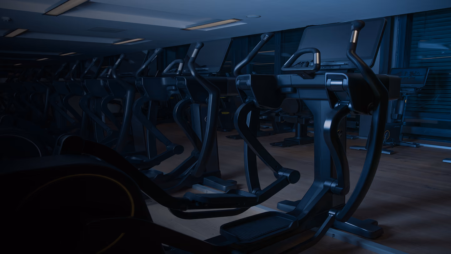Dimly lit gym room with a row of elliptical machines on a wooden floor under fluorescent ceiling lights.