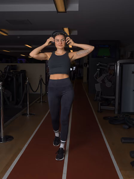 Woman in black workout outfit walking on indoor track in gym adjusting black headphones.