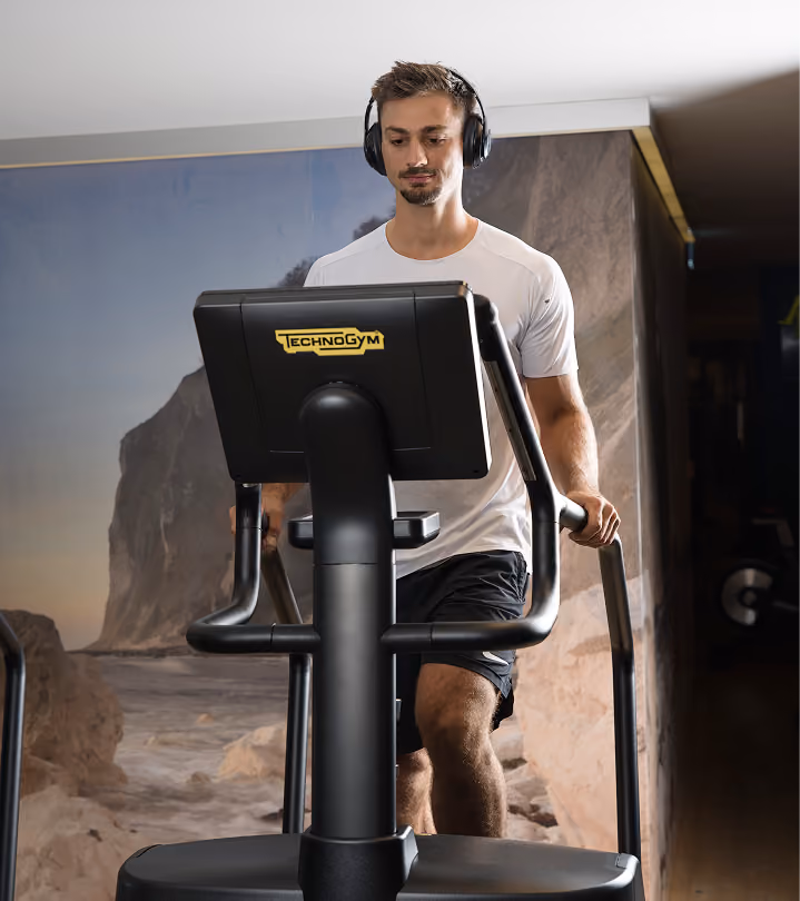 A man wearing headphones and a white t-shirt exercises on a Technogym elliptical machine indoors with a wall mural of a rocky beach in the background.