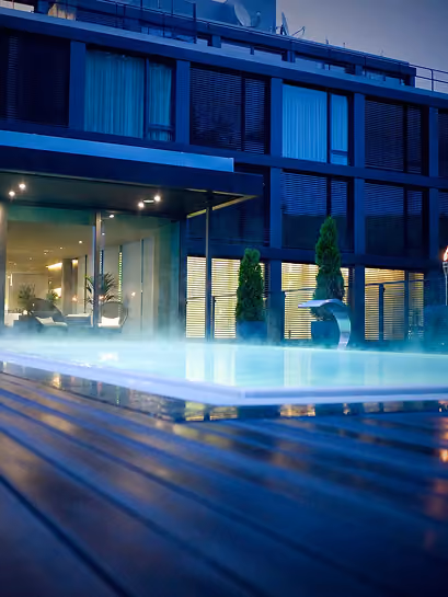 Modern building at dusk with illuminated interior, featuring a swimming pool with steam rising and a wooden pool deck in the foreground.