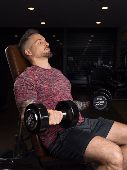 Man with a short beard and tattoos on his left arm seated on a gym bench, lifting two heavy dumbbells while leaning back slightly, wearing a red and gray athletic shirt and black shorts.