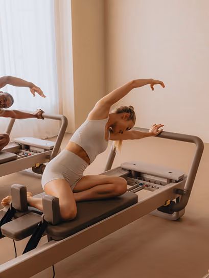 A woman in light gray athletic wear stretches sideways on a Pilates reformer machine inside a room with beige walls and light coming through a window.