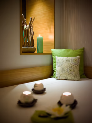 Relaxing spa or wellness room with a cushioned bench featuring green and patterned pillows, a lit green candle, and decorative wooden sculptures on a wall shelf.