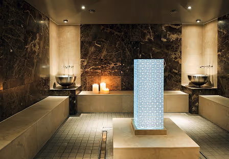 Modern spa room with marble walls, stone seating, a central illuminated decorative pillar, two metal basins on dark shelves, and lit candles near a seating platform.