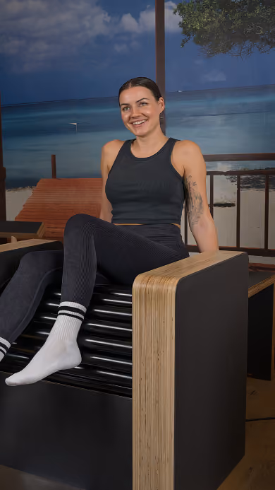 A smiling woman with tied-back dark hair, wearing a black sleeveless top and black leggings, sitting relaxed on a black and wood-accented modern chair with one leg raised, indoors in front of a wall mural depicting a beach scene with blue sky and ocean.