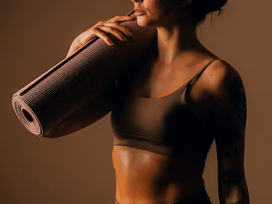 Woman in a dark sports bra holding a rolled yoga mat on her shoulder against a brown background.