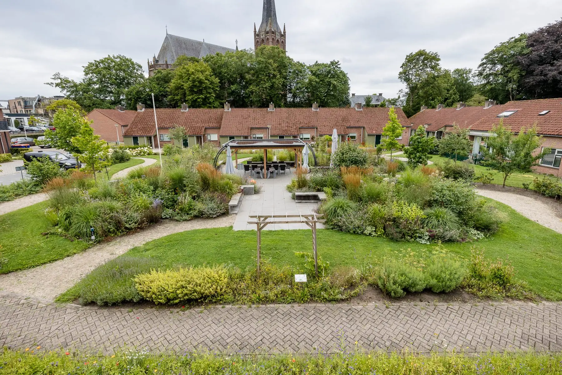 Gemeenschappelijke binnentuin in Raalte met houten overkapping, terras en borders met siergrassen, met de kerktoren op de achtergrond