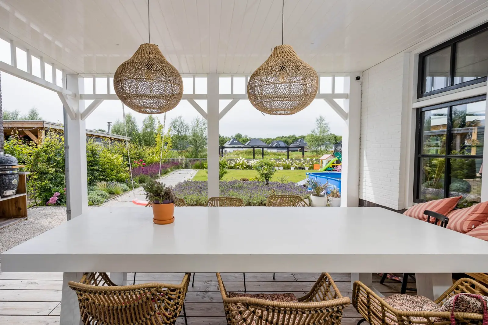 Witte eettafel met rotan stoelen en rotan hanglampen onder de veranda met zicht op de tuin in Lelystad