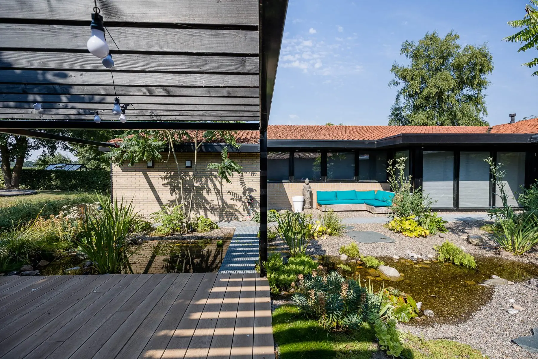 Binnentuin in Enschede met composietvlonder onder een zwarte pergola, een natuurlijk beekje met varens en een loungehoek