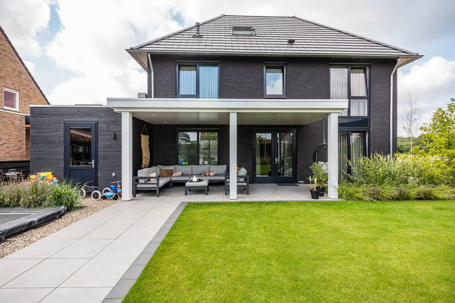 Moderne witte overkapping aan de donkere achtergevel met loungeset, terras van keramische tegels en gazon in Kloosterhaar