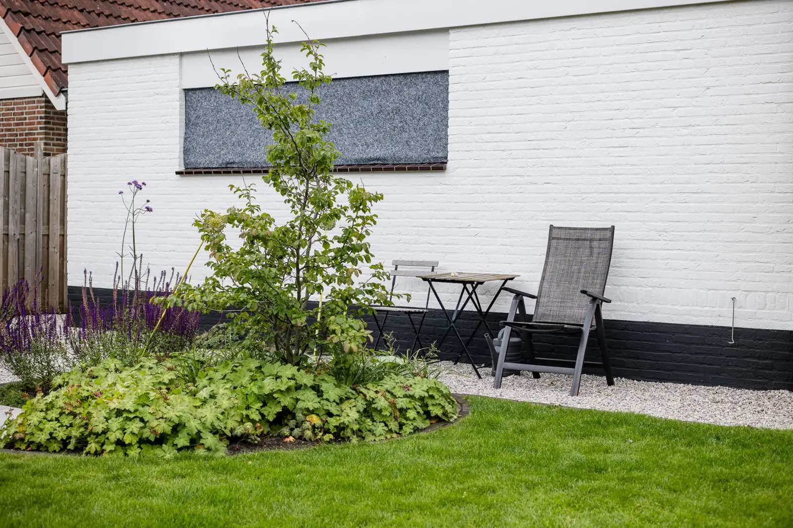 Zitplek op grind tegen de witte gevel naast een ronde border met geranium en salie
