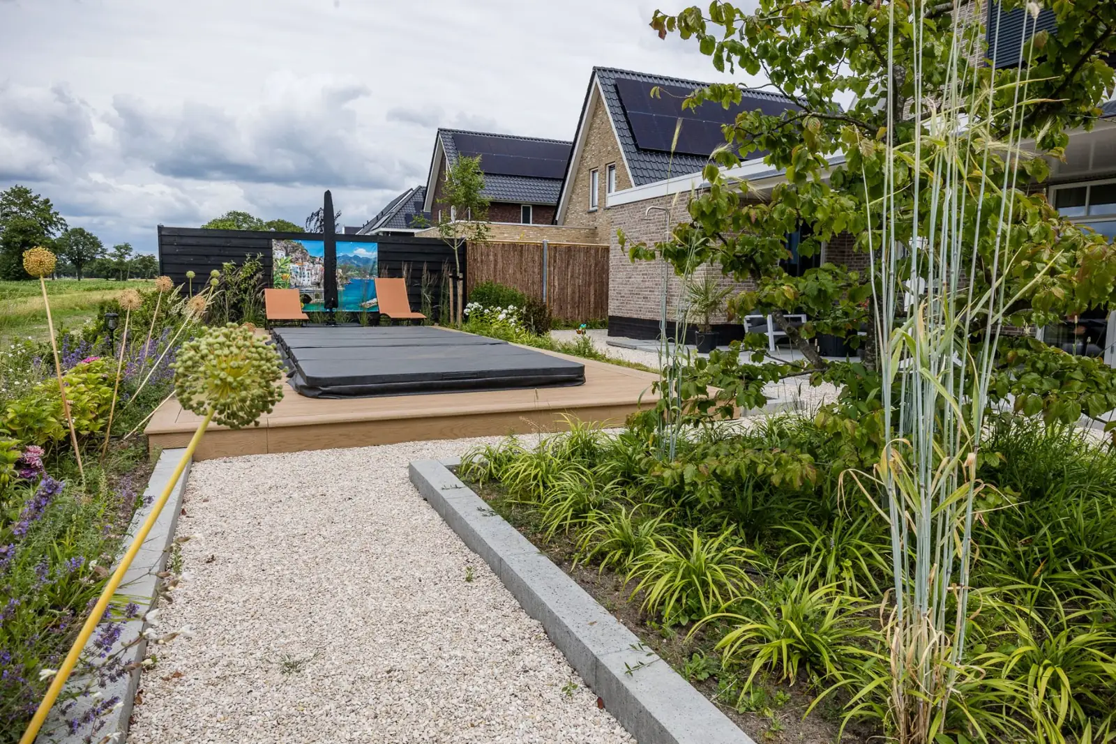Jacuzzi op een verhoogde vlonder langs een grindpad met sierui en kattenkruid in Hardenberg