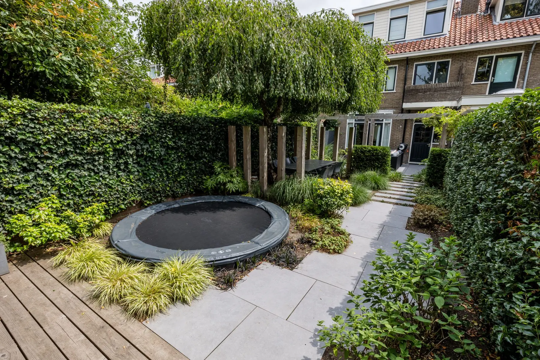 Overzicht van een stadstuin in Zwolle met ingegraven trampoline, houten palen en tegelpad
