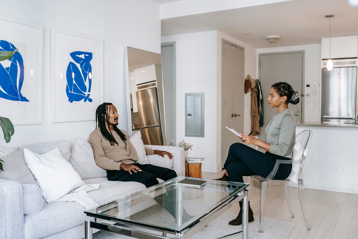 A counselor with a clipboard talks with a man sitting on a sofa in a bright living room.