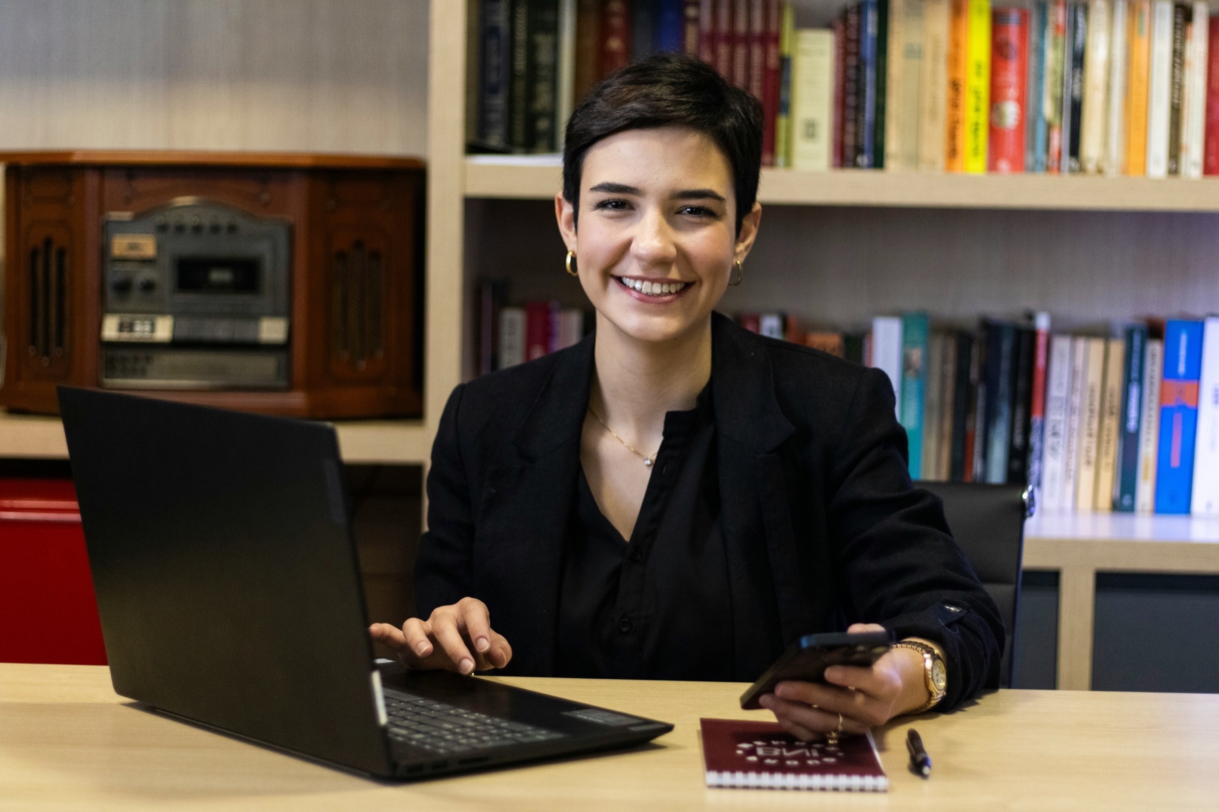 A smiling woman at a desk in front of bookshelves.