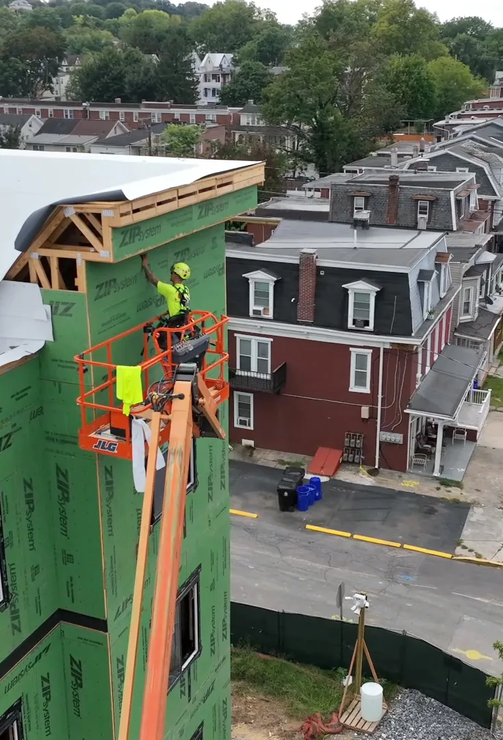 A worker in a lift basket works at the roof edge of the green-sheathed building, above a row of brick houses.