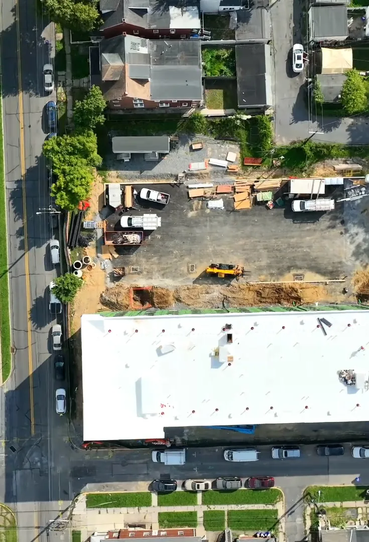 Top-down drone view of the long white roof, the cleared site behind it and the street with parked cars in front.