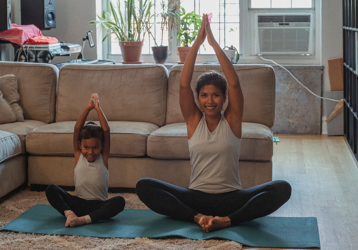 A mother and her young daughter stretch together on a mat in their living room.