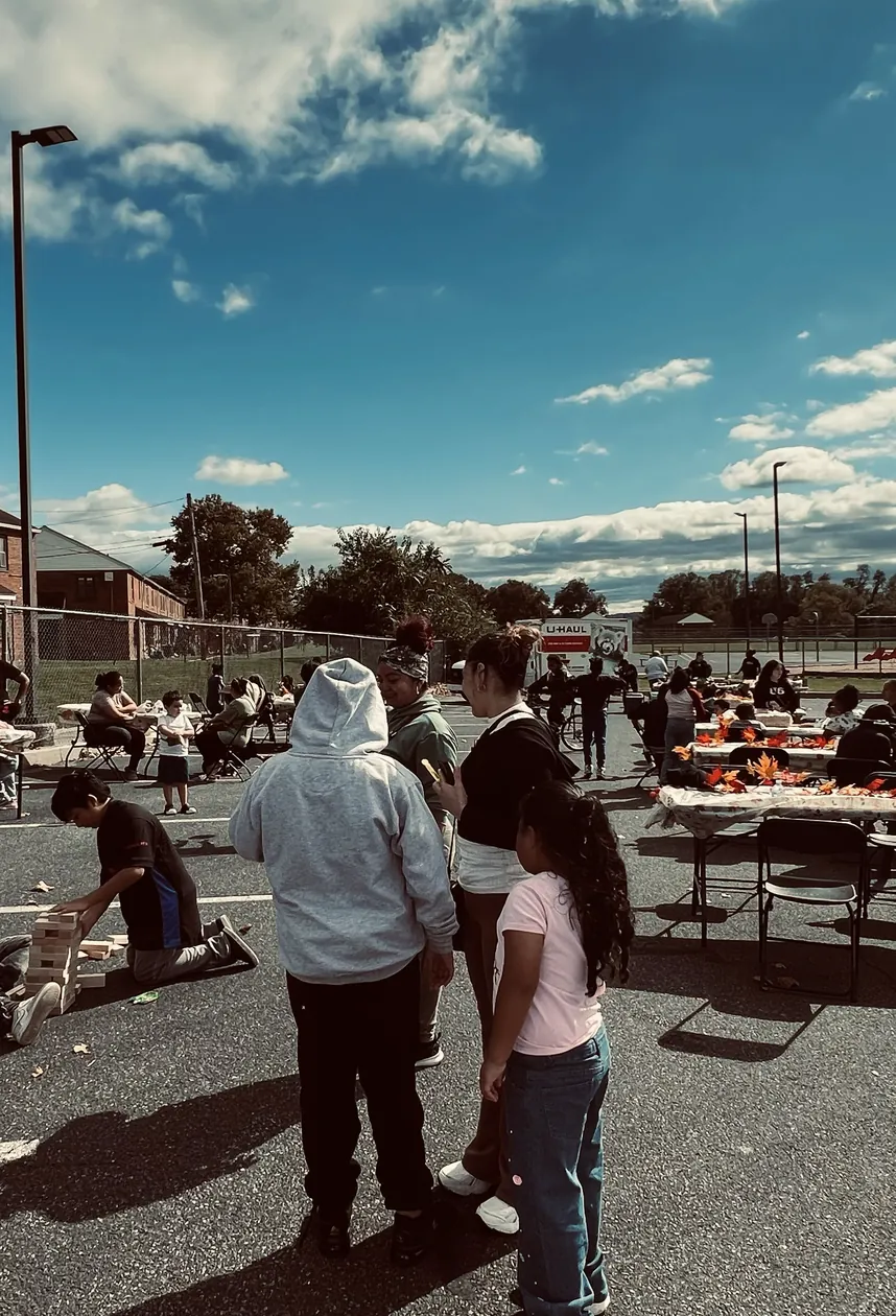 Children and families at tables and games in a parking lot under a blue sky with clouds.